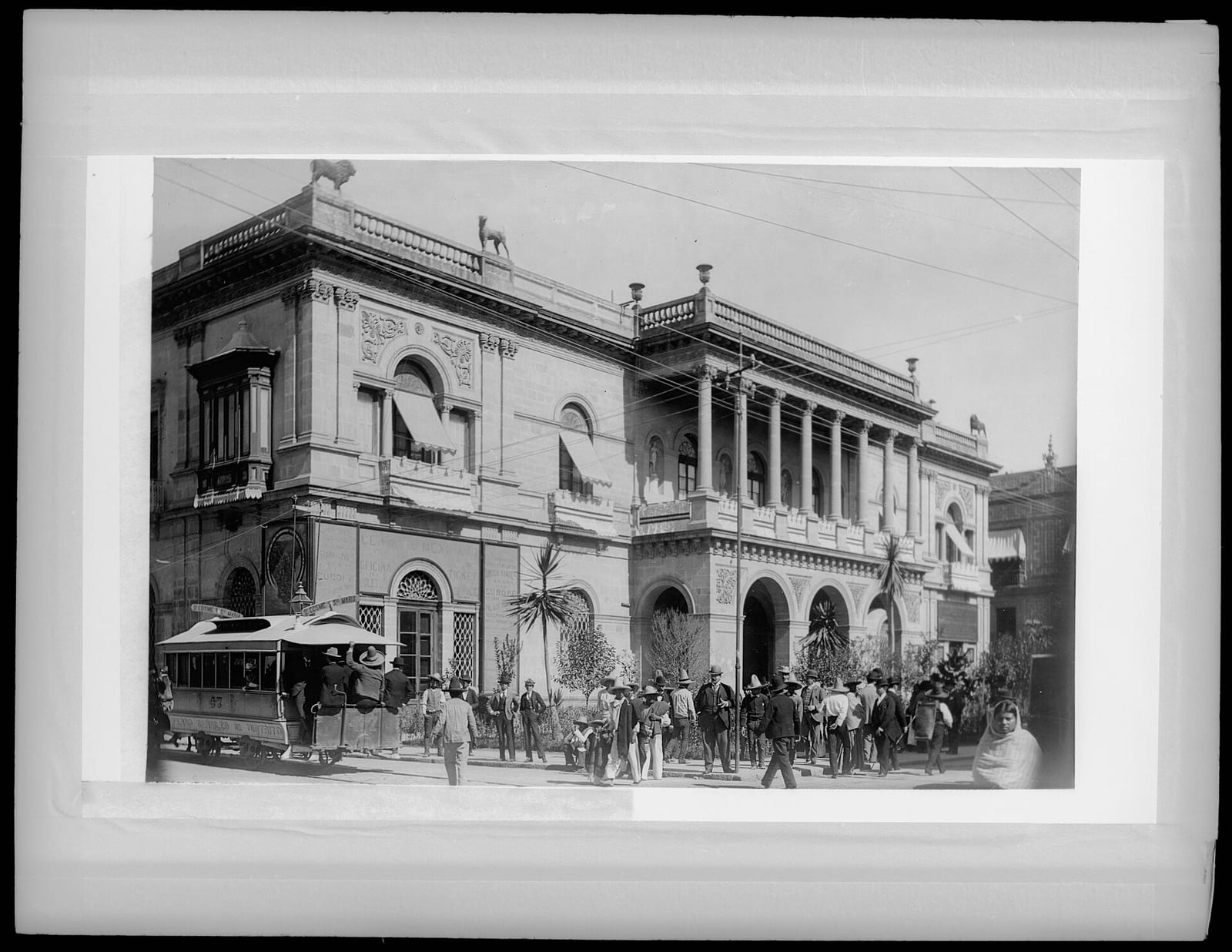 Fotografía de un palacio de dos pisos con balcones y columnas en una esquina de la Ciudad de México, con un tranvía y transeúntes.