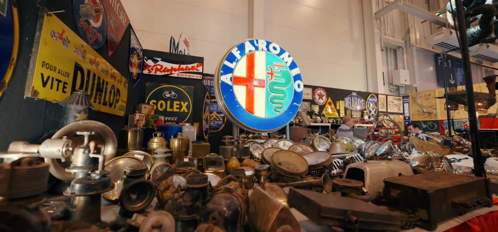 Puesto de automovilia con piezas antiguas, faros, carburadores y un gran letrero de Alfa Romeo.