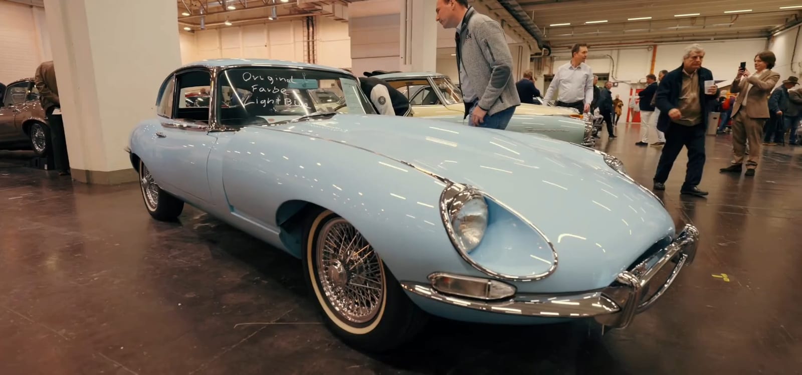 Jaguar E-Type azul cielo exhibido en un pabellón, con visitantes alrededor.