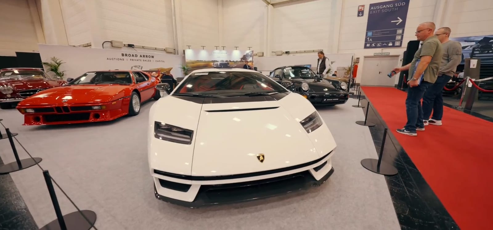 Lamborghini Countach blanco al frente y un BMW M1 rojo detrás, en un módulo con alfombra roja.